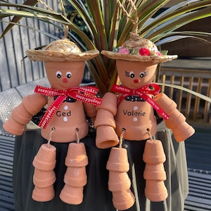 May include: Two terracotta pot dolls with straw hats and painted faces. One doll has a red ribbon that says "Ged" and "Hugs & Kisses." The other doll has a ribbon that says "Valerie" and "I Love You."
