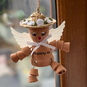 May include: A terracotta angel figurine with white wings, a straw hat with white flowers, and a white ribbon. The angel is holding a small terracotta pot with the text "Nanny xx" written on it.