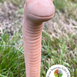 May include: A terracotta worm garden stake with a smiling face and black eyes. The worm is standing upright in a grassy area. The stake is a decorative garden ornament. The image includes a logo for Four Seasons Gifts UK.