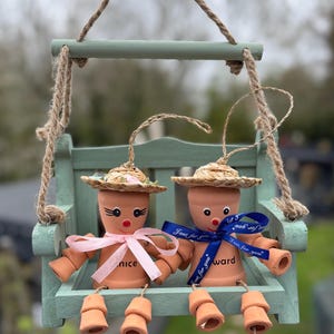 May include: Two terracotta pot figures with straw hats and painted faces sit on a small, mint-green wooden swing. Each figure has a ribbon bow, one pink and one blue, with text. The swing hangs from jute rope.