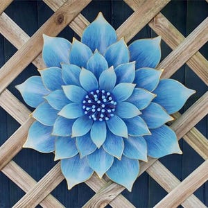 Puede incluir: Una gran flor de metal azul claro con ribetes dorados, montada sobre un enrejado de madera. La flor tiene m&uacute;ltiples capas de p&eacute;talos y un centro detallado. El fondo es una superficie azul oscuro. Un adorno decorativo para el jard&iacute;n.