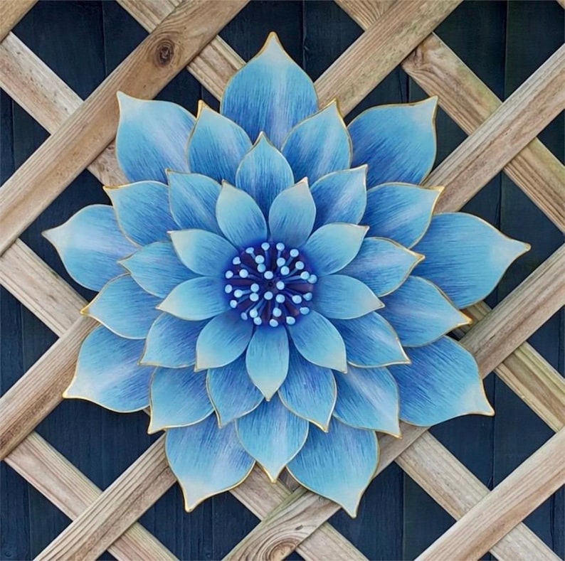 Puede incluir: Una gran flor de metal azul claro con ribetes dorados, montada sobre un enrejado de madera. La flor tiene m&uacute;ltiples capas de p&eacute;talos y un centro detallado. El fondo es una superficie azul oscuro. Un adorno decorativo para el jard&iacute;n.