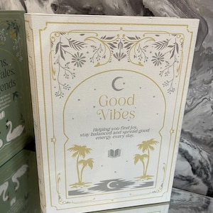 May include: A white book with gold and gray accents, featuring the words "Good Vibes" in elegant script. The cover includes illustrations of palm trees, a crescent moon, and floral designs. The text reads: "Helping you find joy, stay balanced and spread good energy every day."