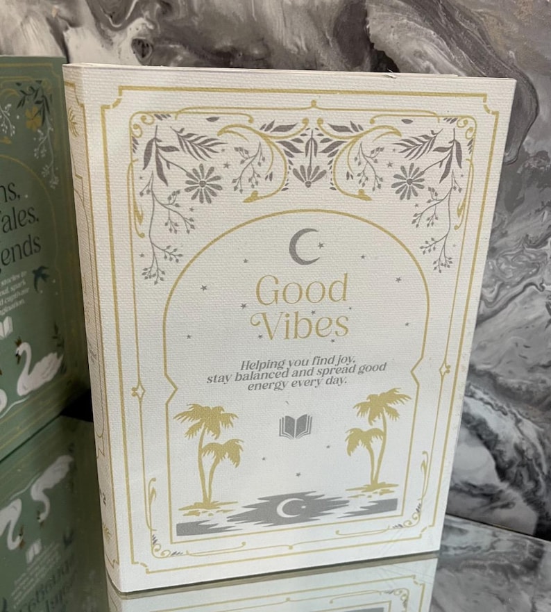 May include: A white book with gold and gray accents, featuring the words "Good Vibes" in elegant script. The cover includes illustrations of palm trees, a crescent moon, and floral designs. The text reads: "Helping you find joy, stay balanced and spread good energy every day."