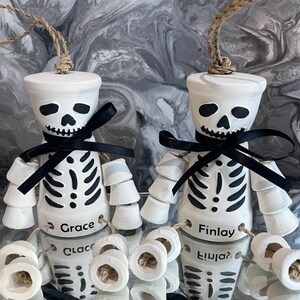 May include: Two white ceramic skeleton ornaments with black ribbon bows and black painted details. The ornaments are labelled "Grace" and "Finlay".