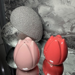 May include: A pair of ceramic salt and pepper shakers shaped like tulip flowers. One shaker is light pink, the other is red. A silver decorative sphere and a silver egg-shaped object are in the background.