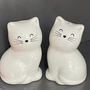 May include: A pair of white ceramic cat-shaped salt and pepper shakers. Each shaker features a smiling face with black whiskers and a small hole on top. The ears and inner ears are a pale pink colour.