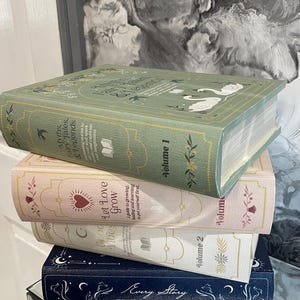 May include: A stack of decorative books in various colors. The top book is sage green with the title "Myths, Fairy Tales, & Legends." Below are books in pink, white, and navy blue, each with unique titles and designs. The books are stacked on a reflective surface.
