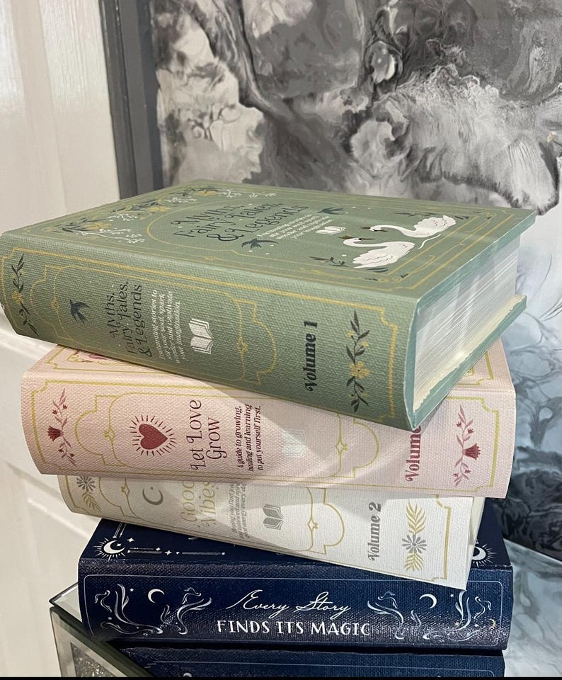 May include: A stack of decorative books in various colors. The top book is sage green with the title "Myths, Fairy Tales, & Legends." Below are books in pink, white, and navy blue, each with unique titles and designs. The books are stacked on a reflective surface.