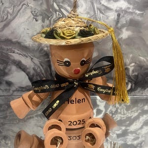May include: A terracotta graduation ornament shaped like a doll, wearing a straw hat decorated with green and black roses. A black ribbon with "Congrats Grad" is tied around the doll. The doll has the name "Helen" and the year "2025" written on it.