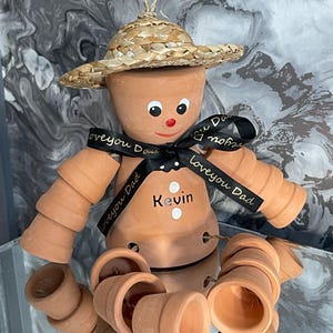 May include: A terracotta pot figure wearing a straw hat, with a black ribbon reading "Love you Dad" and the name "Kevin". The figure has a painted face with a red nose and is surrounded by small terracotta pots.