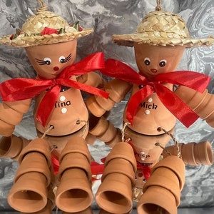 May include: Two terracotta pot people with straw hats and red ribbon bows. The pot people are decorated with painted faces and the names "Tina" and "Mick". Each pot person has a stack of smaller terracotta pots hanging from their arms.