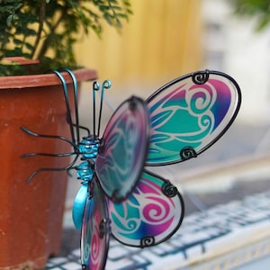 May include: A colourful metal butterfly garden stake with blue, purple and green iridescent wings. The butterfly is perched on a potted plant.
