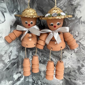 May include: Two terracotta pot people with straw hats and white ribbon bows. The pot people are decorated with painted faces and the names "Lain" and "Maui".