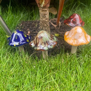 K&ouml;nnte beinhalten: Eine Sammlung von Keramik-Gartenornamenten in Pilzform. Die Pilze sind in verschiedenen Farben gehalten, darunter Blau mit gelben Punkten, Gr&uuml;n, Orange und Rot. Sie stehen im gr&uuml;nen Gras und sind eine dekorative Idee.