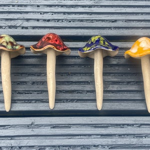 K&ouml;nnte beinhalten: Vier Keramik-Pilzfiguren mit unterschiedlichen Hut-Farben: Braun und Gr&uuml;n, Rot, Blau und Gelb. Jeder Pilz hat einen cremefarbenen Stiel. Die Pilze sind auf einer grauen Holzoberfl&auml;che angeordnet.