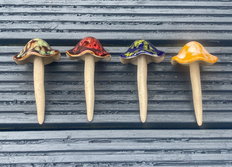 K&ouml;nnte beinhalten: Vier Keramik-Pilzfiguren mit unterschiedlichen Hut-Farben: Braun und Gr&uuml;n, Rot, Blau und Gelb. Jeder Pilz hat einen cremefarbenen Stiel. Die Pilze sind auf einer grauen Holzoberfl&auml;che angeordnet.