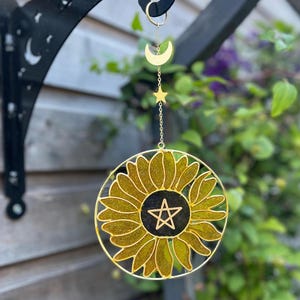 May include: A gold-coloured suncatcher with a sunflower design, a black centre, and a pentagram. It hangs from a gold chain with a crescent moon and star charm. The background includes a black metal bracket and green foliage.
