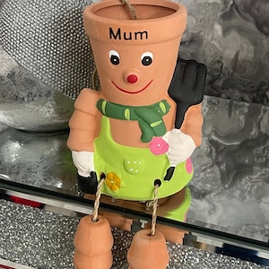 May include: A terracotta planter shaped like a person with a green apron, black gardening gloves, and a green scarf. The planter has a smiling face and the word "Mum" written on the front.