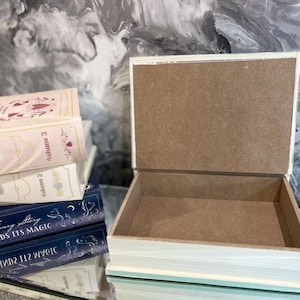 May include: An open, rectangular wooden box with a light blue and white painted exterior. Several books are stacked nearby, with titles including "Let Love Grow" and "Every Story Finds Its Magic."