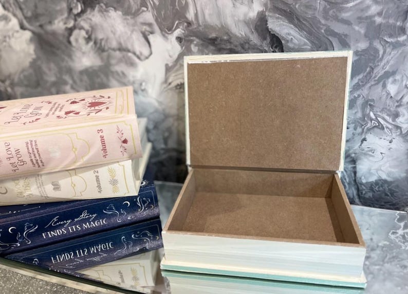 May include: An open, rectangular wooden box with a light blue and white painted exterior. Several books are stacked nearby, with titles including "Let Love Grow" and "Every Story Finds Its Magic."