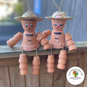 May include: Two terracotta pot people with painted faces and straw hats. The pot people are labelled "Mr" and "Mrs".
