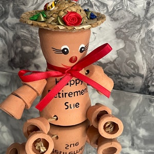 May include: A terracotta figurine of a person wearing a straw hat with red, green and blue accents. The figurine has a red ribbon around its neck and the words "Happy Retirement Sue" written on its torso. The figurine has multiple terracotta pots attached to its legs.