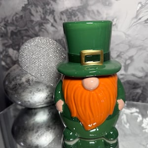 May include: A ceramic gnome figurine with a green top hat and body, an orange beard, and a gold buckle. The gnome has a round, pink nose and is a festive decoration.