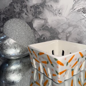 May include: A white ceramic square berry bowl with orange carrot and pink flower designs. The bowl has cut-out holes and is reflected on a mirrored surface. A silver decorative object is in the background.