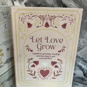 May include: A pink book with the words "Let Love Grow" in elegant script. The cover features floral and heart illustrations in burgundy and green. The text below reads "A guide to growing, healing and learning to put yourself first."