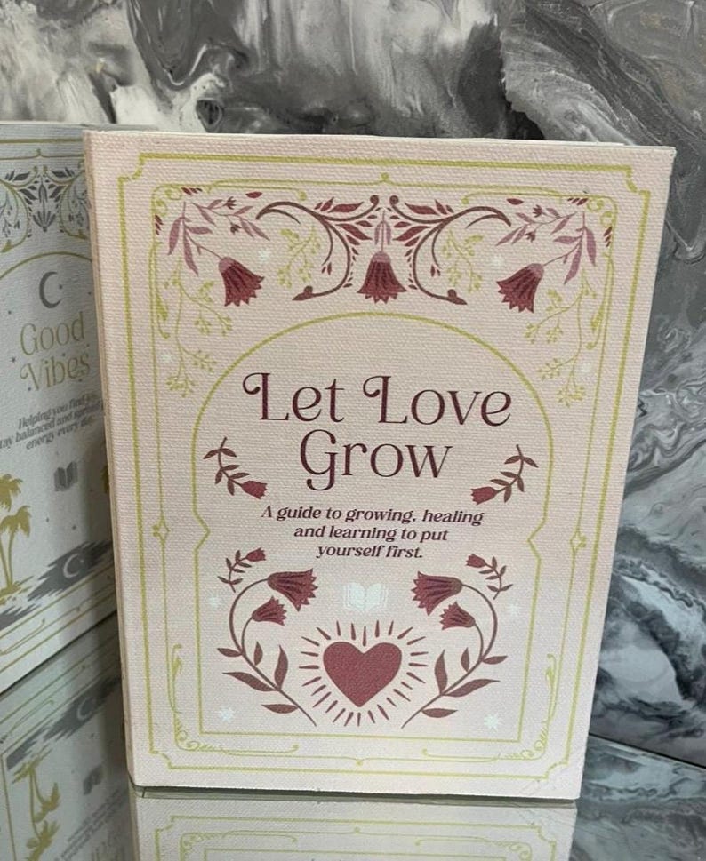 May include: A pink book with the words "Let Love Grow" in elegant script. The cover features floral and heart illustrations in burgundy and green. The text below reads "A guide to growing, healing and learning to put yourself first."