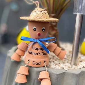 May include: A terracotta figurine of a person wearing a straw hat and a blue ribbon. The figurine has a painted face and the words "Happy Father's Day Dad" are written on the front.
