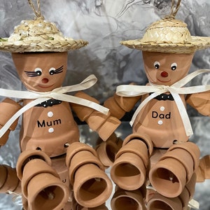 May include: Two terracotta pot people with straw hats and white ribbons. The pot person on the left has the word "Mum" written on it. The pot person on the right has the word "Dad" written on it.