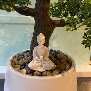 May include: A white ceramic Buddha figurine sitting in a white pot with small grey stones and a green artificial bonsai tree.