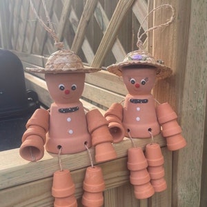 May include: Two terracotta pot people with painted faces and straw hats. The figures have arms and legs made of stacked terracotta pots.