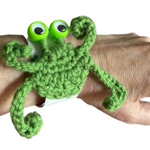 May include: Green crocheted crab bracelet with googly eyes. The crab is attached to a white elastic band.