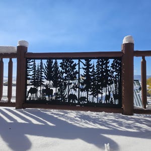 May include: A decorative metal fence panel with a forest scene cut-out. The panel features silhouettes of trees, moose, bears, and deer. The fence is part of a wooden deck covered in snow, with a clear blue sky in the background.