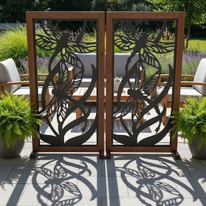 May include: Two wooden outdoor privacy screens with a dark metal dragonfly design. The screens are framed in brown wood and stand on a patio. The dragonfly design casts a shadow on the ground.