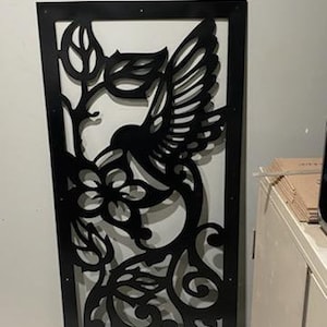 May include: Black metal wall decor featuring a hummingbird in flight with intricate floral details. The design is cut out of the metal, creating a delicate and airy look.