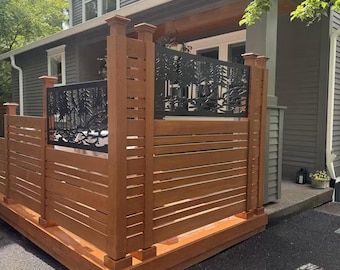 May include: A wooden privacy screen featuring horizontal slats and black metal panels with a laser-cut forest silhouette design. The structure is built on a raised wooden platform, positioned outdoors in front of a gray house with white trim.