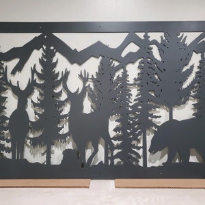 May include: A rectangular black metal wall art panel featuring a laser-cut silhouette of a forest landscape. The design includes two deer standing among tall pine trees, a bear walking on the right side, and mountain peaks in the background.