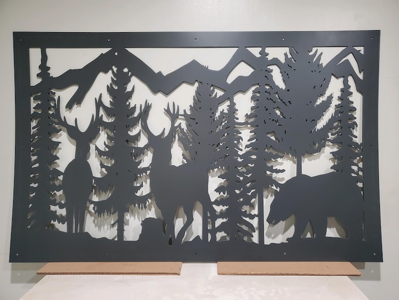 May include: A rectangular black metal wall art panel featuring a laser-cut silhouette of a forest landscape. The design includes two deer standing among tall pine trees, a bear walking on the right side, and mountain peaks in the background.