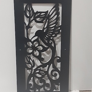 May include: Black metal wall decor featuring a hummingbird in flight with intricate floral details. The hummingbird is depicted with outstretched wings and a long beak, hovering near a stylized flower.