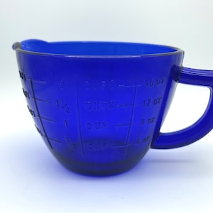 May include: A cobalt blue glass measuring cup with a handle. The cup has measurement markings for cups and ounces. The cup is designed to measure up to 2 cups or 16 ounces.
