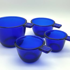 May include: Set of four blue glass measuring cups with handles. The cups are graduated in size, with the largest cup being the tallest.