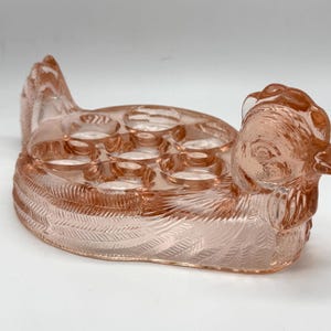 May include: A translucent pink pressed glass egg holder shaped like a sitting hen. The piece features a detailed molded feather pattern across the body and wings, with a raised head and tail. The top surface contains six circular indentations.