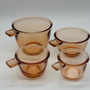 May include: Set of four amber-colored glass measuring cups. Each cup has a handle with measurement markings. The cups are nested together, with the largest cup in the back and the smallest in the front. The cups are made of clear glass.