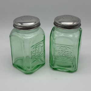 May include: A pair of vintage green glass salt and pepper shakers. Each shaker has a square shape with rounded corners and a silver metal lid. The word "SALT" is embossed on one shaker, and "PEPPER" on the other.