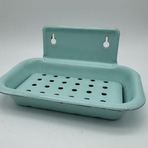 May include: A vintage-style, light blue enamel soap dish with a rectangular shape and a perforated base. The dish has a backplate with two holes for mounting. The edges are trimmed with black. A classic bathroom accessory.