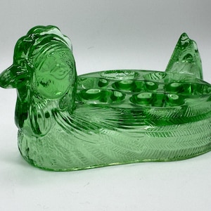 May include: Green glass hen-shaped egg holder. The decorative piece features a detailed hen design with a head, tail, and space for eggs. The egg holder is made of green glass and has a textured surface.